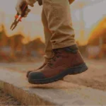 Trabajador en obra de pie sobre concreto con botas ergonómicas, mostrando la suela acolchada mientras sostiene herramientas, con fondo desenfocado de construcción.