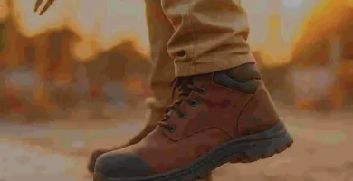 Trabajador en obra de pie sobre concreto con botas ergonómicas, mostrando la suela acolchada mientras sostiene herramientas, con fondo desenfocado de construcción.