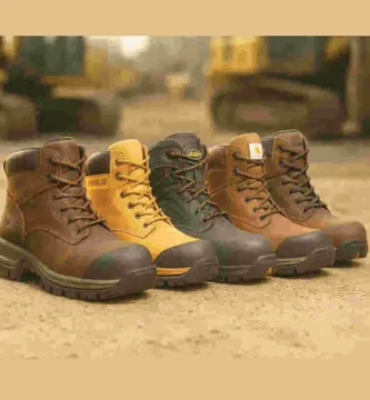 Cinco botas de trabajo alineadas sobre suelo de construcción, cada una con diferentes colores y tipos de punta, con maquinaria industrial desenfocada al fondo.