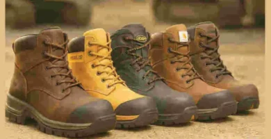 Cinco botas de trabajo alineadas sobre suelo de construcción, cada una con diferentes colores y tipos de punta, con maquinaria industrial desenfocada al fondo.